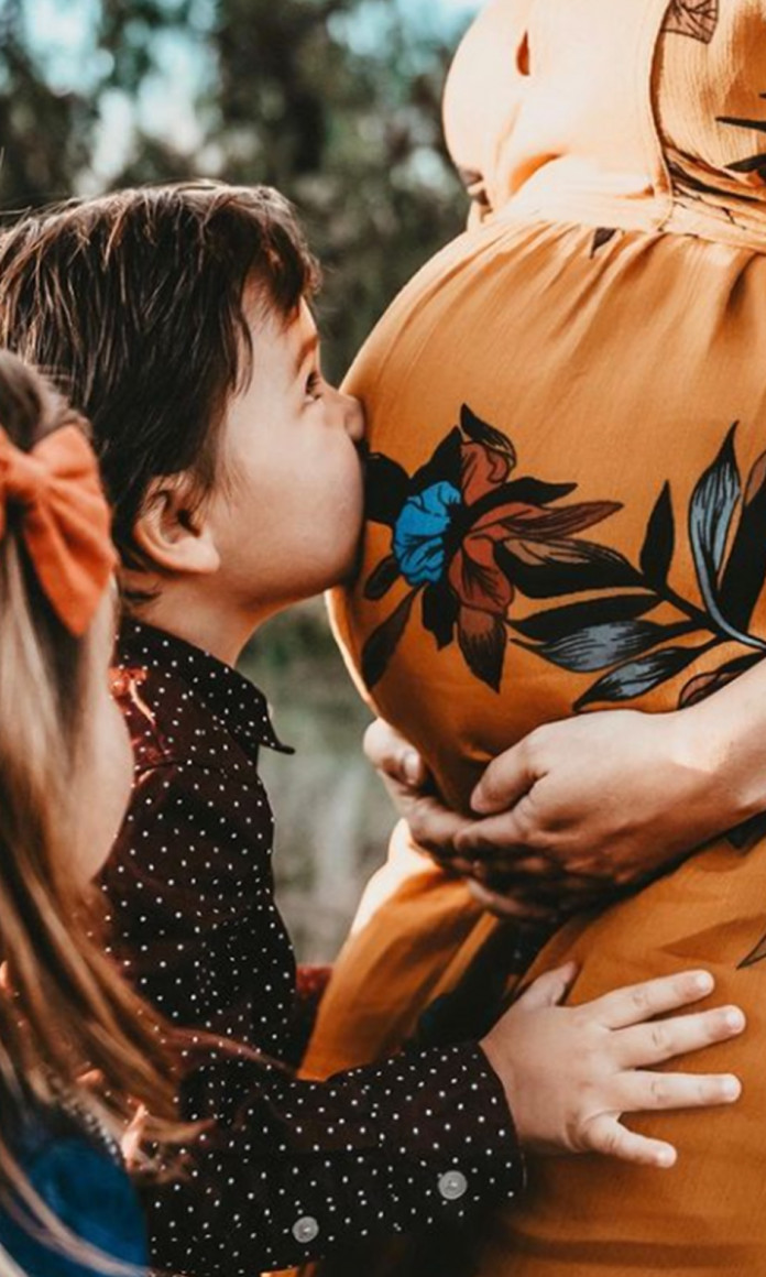 Crianças beija a barriga da mãe, que está em pé e usa um vestido laranja