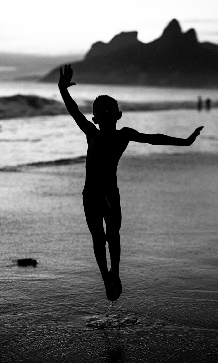 Garoto salta no ar na beira do mar com o Morro dos Irmãos ao fundo, no Arpoador, Rio de Janeiro