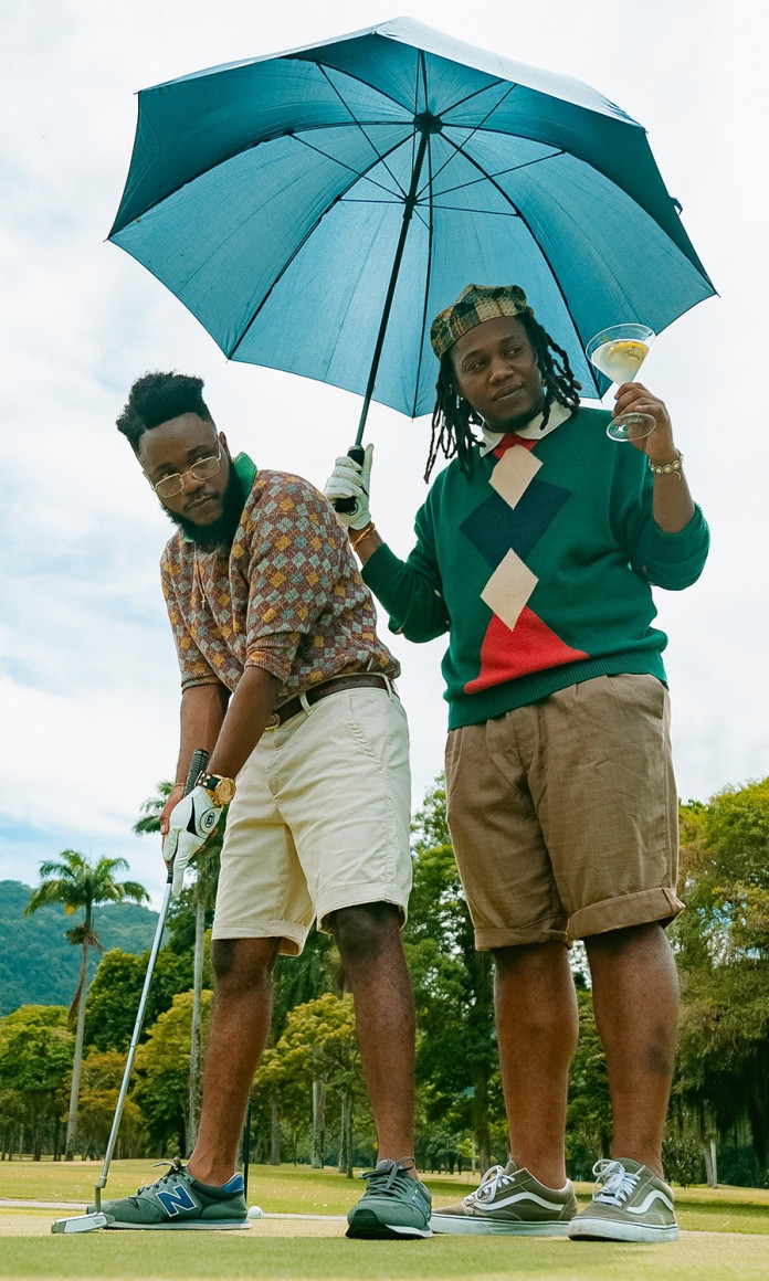 Shuna e Gian Pedro, do duo carioca Yoùn, em cena do clipe, aparecem num campo de golpe segurando um guarda-chuva e um drink