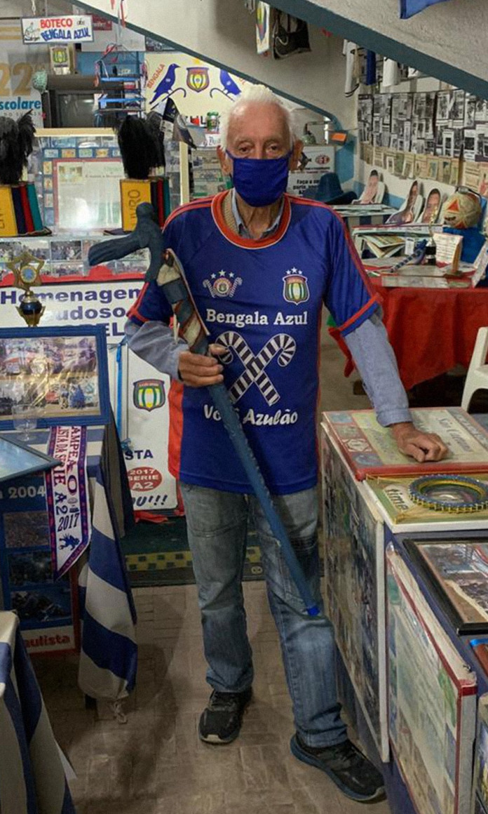 Com uma bengala azul na mão, um senhor posa numa sala cheia de recortes de jornal e trofeus com a camisa da torcida Bengala Azul, do clube Sao Caetano