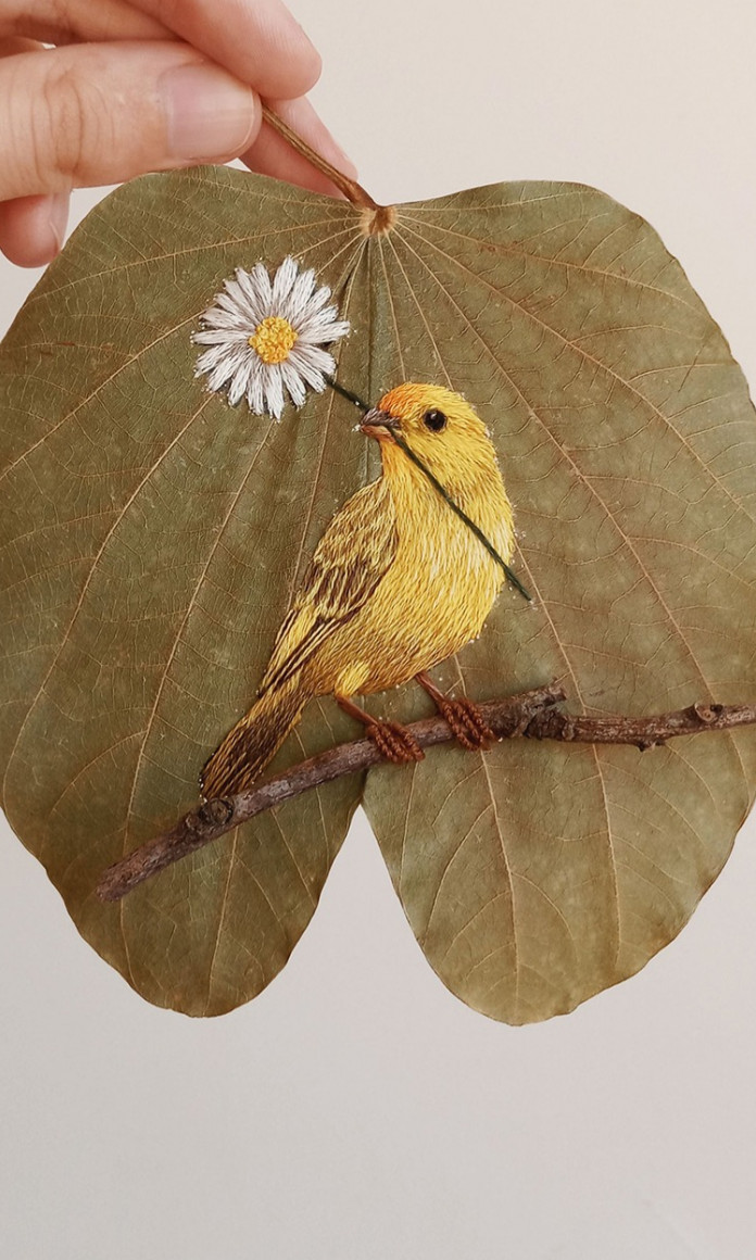 Folha seca com um passarinho amarelo com uma margarida no bico bordado com linha