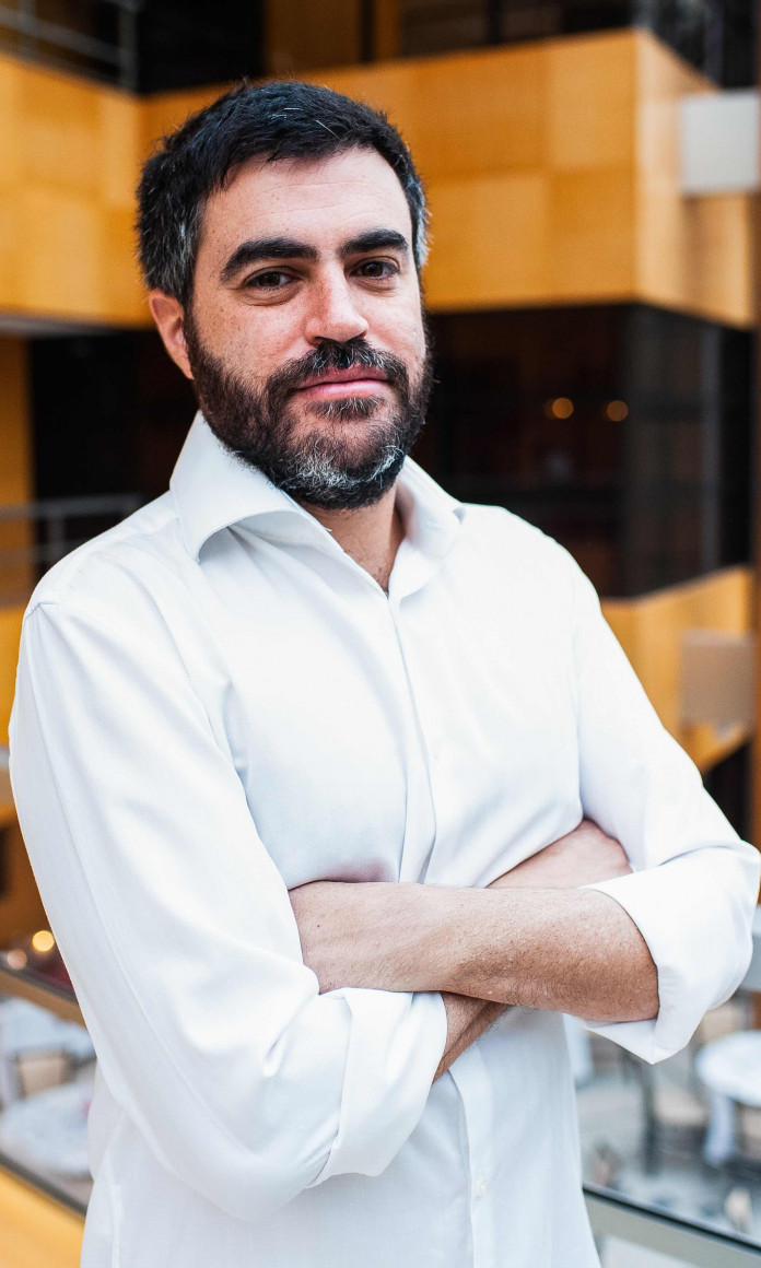 Homem branco com cabelo e barba grisalha veste camisa branca e está de braços cruzados