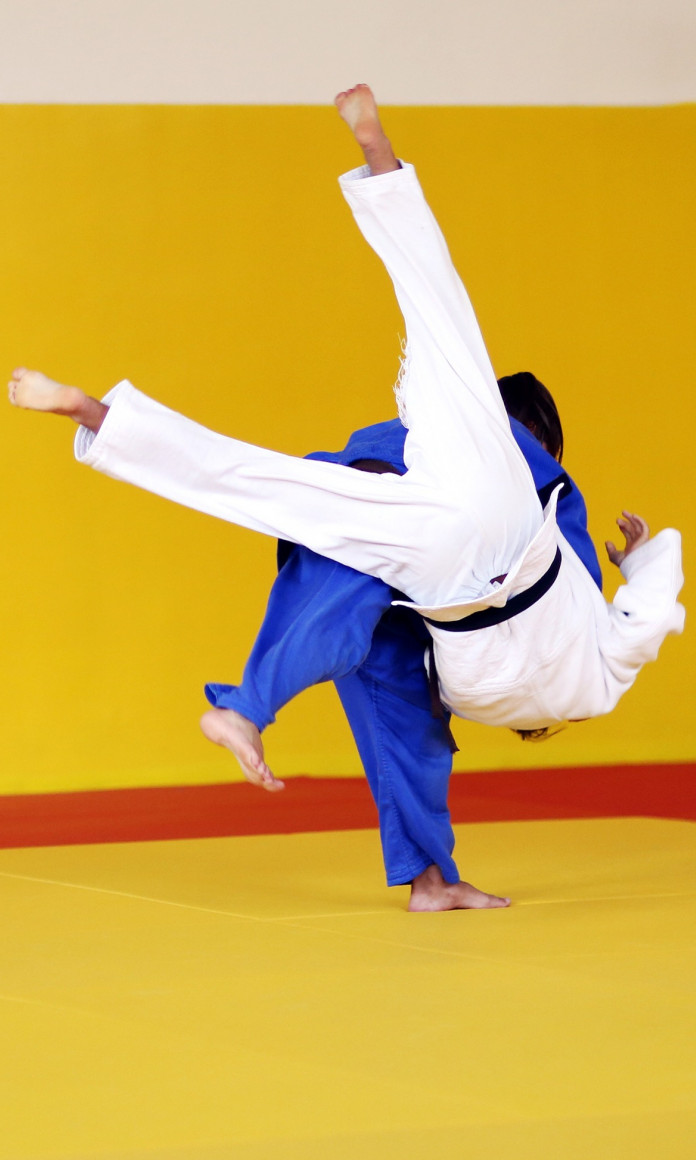 Num tatame amarelo com uma listra vermelha, lutador de uniforme azul levanta o lutador de uniforme branco no ar com um golpe