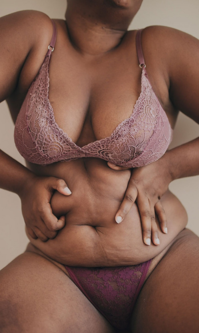 Mulher gorda celebra seu corpo com as mãos na barriga vestindo uma lingerie roxa