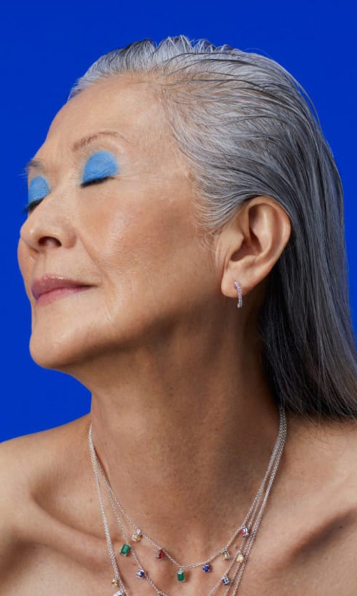 Mulher de 70 anos com cabelos brancos compridos está de olhos fechados e maquiagem azul