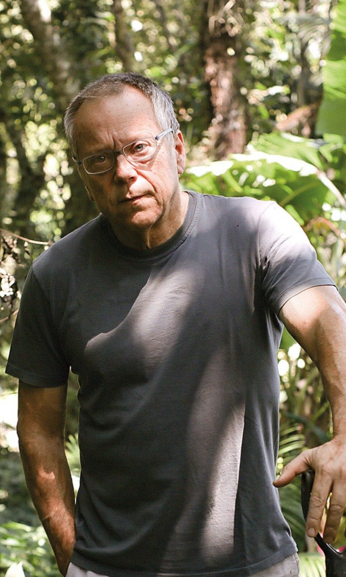 Homem branco de cabelos brancos e óculos e camiseta cinza segura uma pá com um matagal atrás