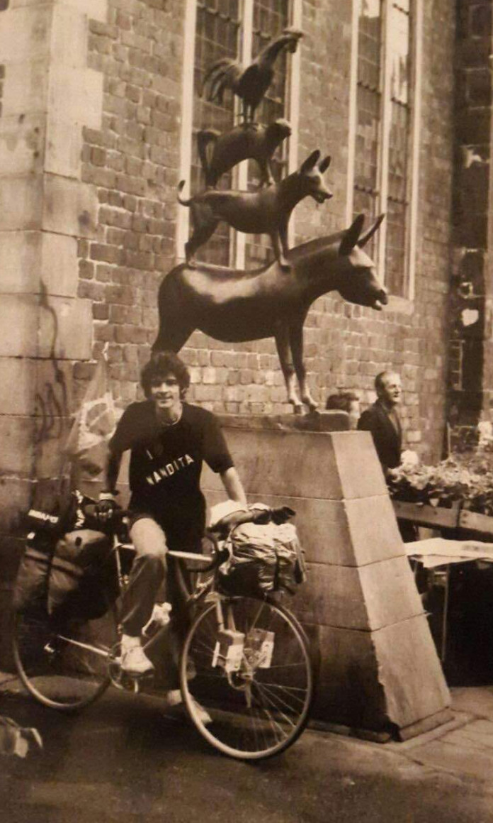 Homem de camiseta preta e cabelos encaracolados está apoiado em uma bicicleta em frente a uma escultura de animais