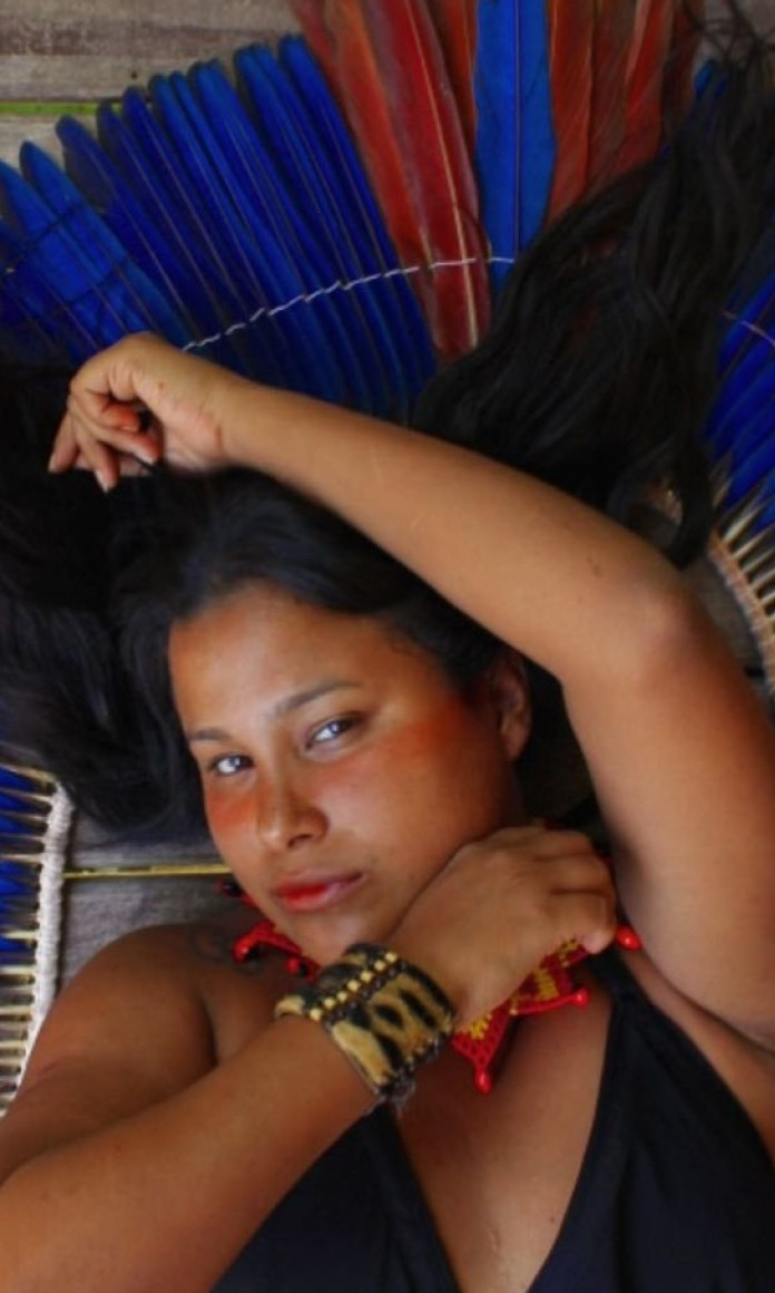 Mulher indígena está deitada com os cabelos castanhos espalhados pelo chão e um cocar azul atrás