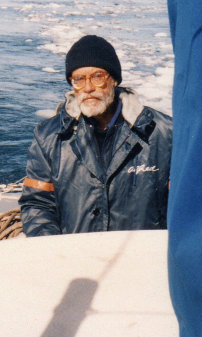 Homem de barba branca, oculos de grau e gorro preto está num barco com o mar atrás