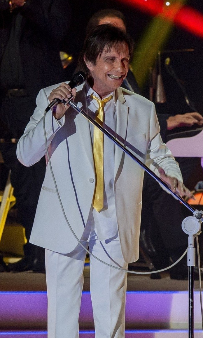 Roberto Carlos tem os cabelos compridos e veste um terno cinza com uma gravata amarela segurando um microfone em pé