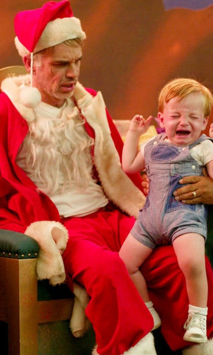 Duende de Natal segura um bebê chorando ao lado de um homem vestido de Papai Noel