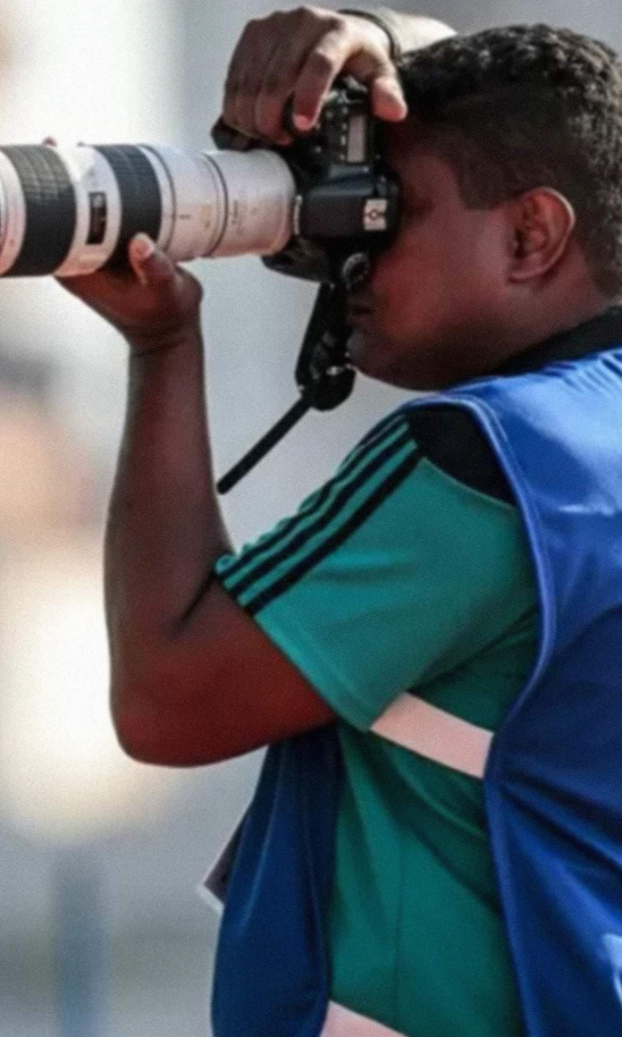 Homem vestido com colete de imprensa segurando uma camera na frente do rosto com uma lente enorme