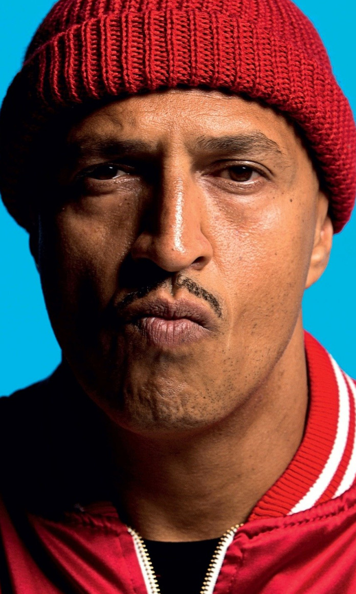 Mano Brown, um homem negro, de bigode, usa gorro e casaco vermelho