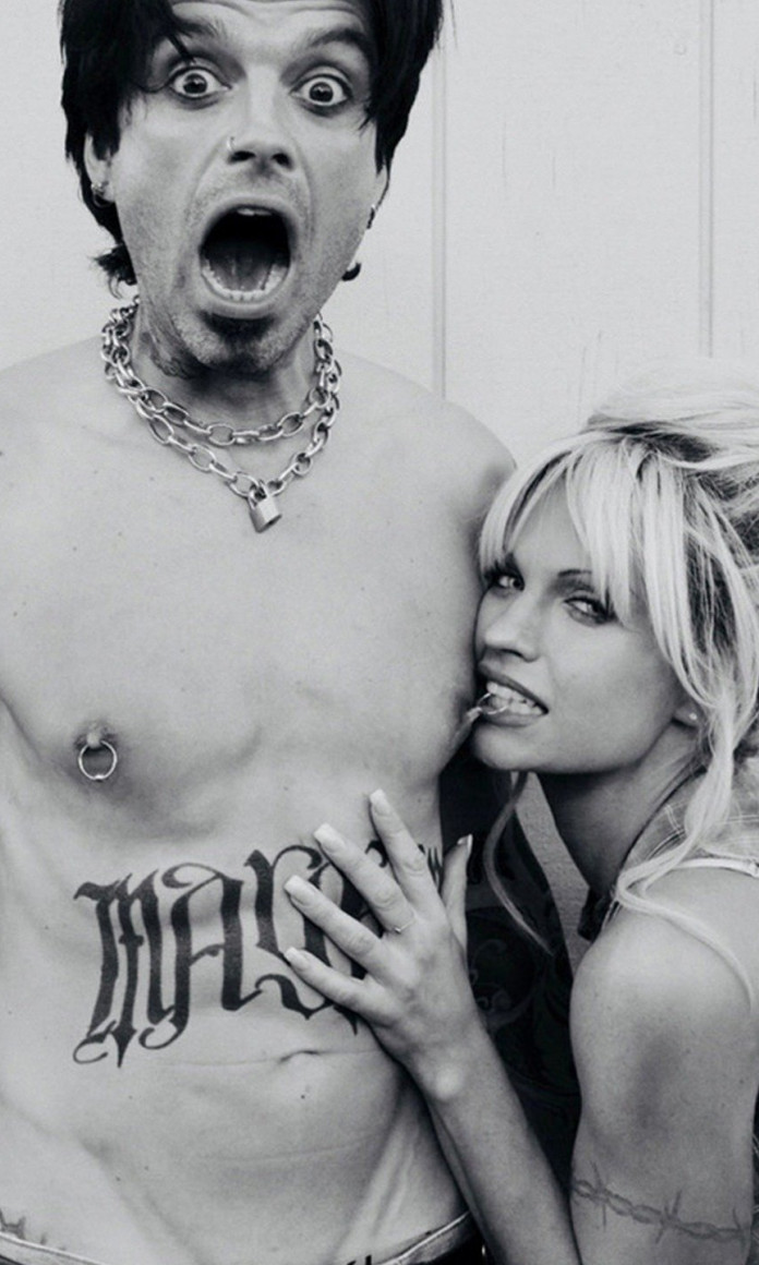 Tommy Lee, uma homem branco e tatuado, aparece sem camisa enquanto Pamela Anderson, loira de cabelos lisos, morde o piercing em seu mamilo