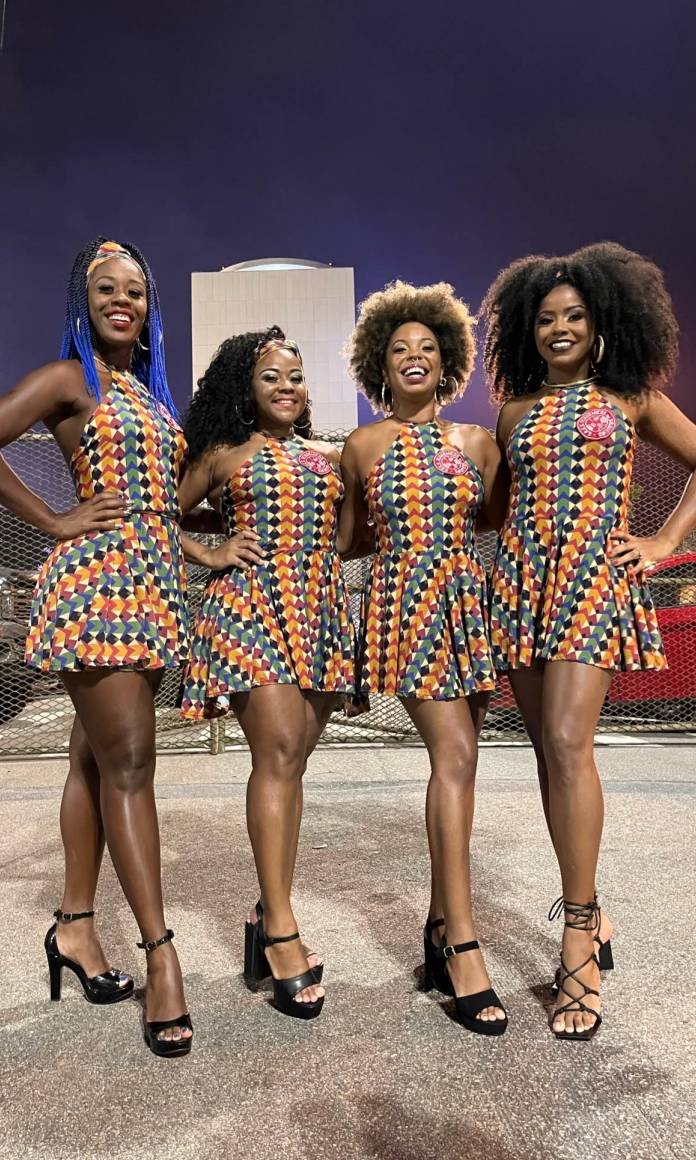 Quatro mulheres negras na avenida do Carnaval usando vestidos iguais de bolinhas laranjas e azuis