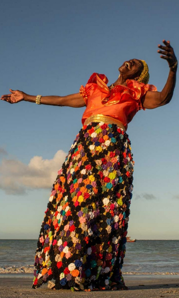 Lia de Itamaraca, uma mulher negra, danca com os bracos para o alto numa praia vestindo uma roupa colorida