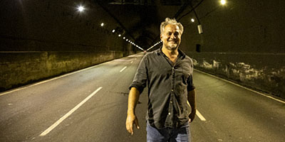 Breno Silveira andando em um túnel iluminado, sem carros, a noite, vestindo calça jeans e camisa, com sorriso no rosto