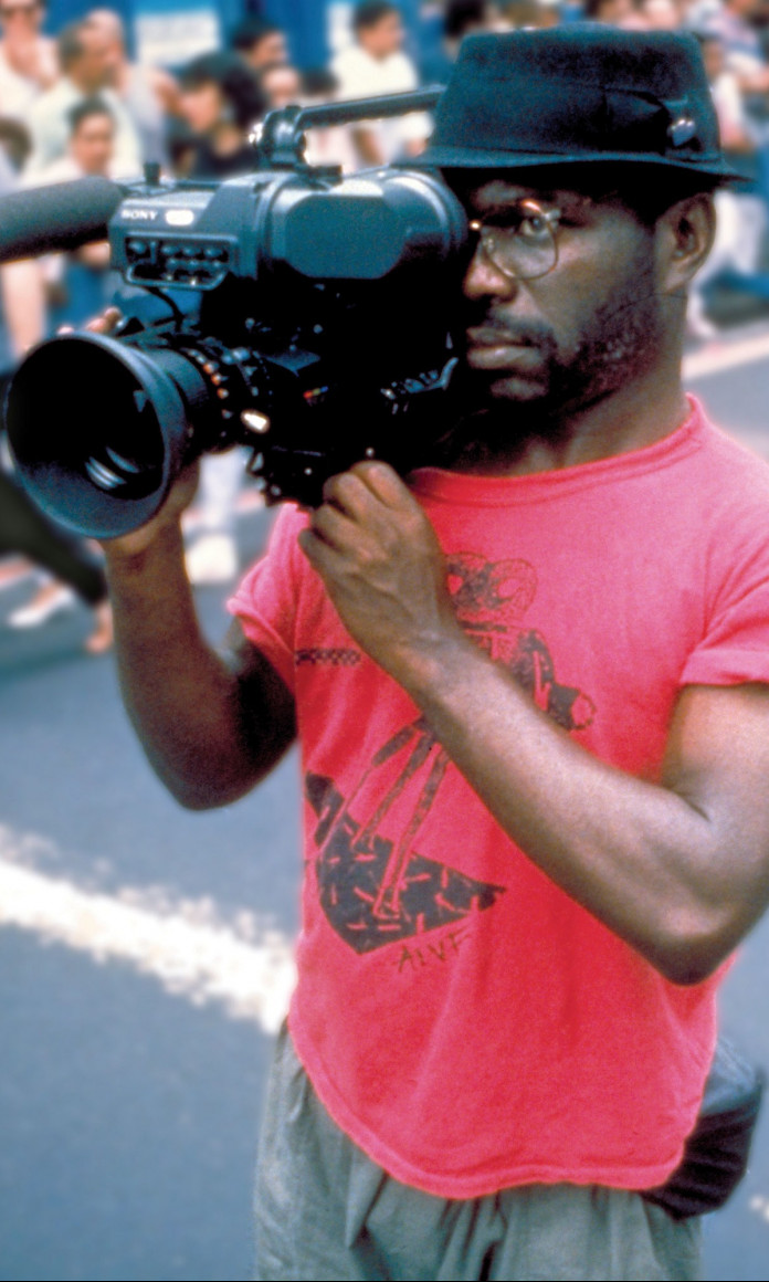 Homem negro com chapeu preto, blusa rosa e uma camera filmografica antiga nos olhos