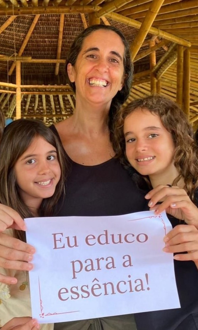 Mulher branca cercada de crianças segurando uma placa: eu educado para a essência