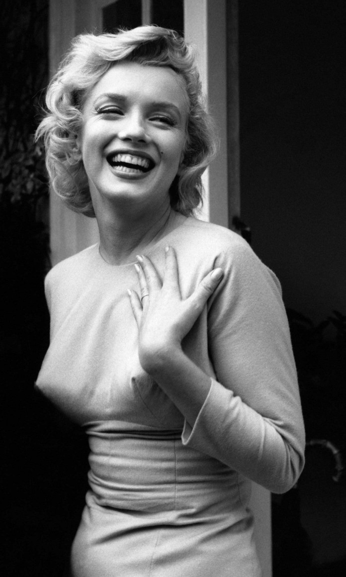 Marilyn Monroe, uma mulher loira, de cabelos curtos e roupa justa, sorri com o braço no colo