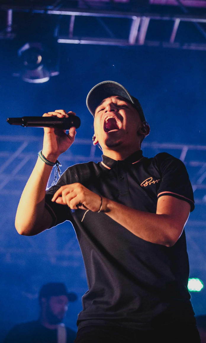 O cantor Joao Gomes, de camiseta e bone pretos, segura o microfone num palco iluminado